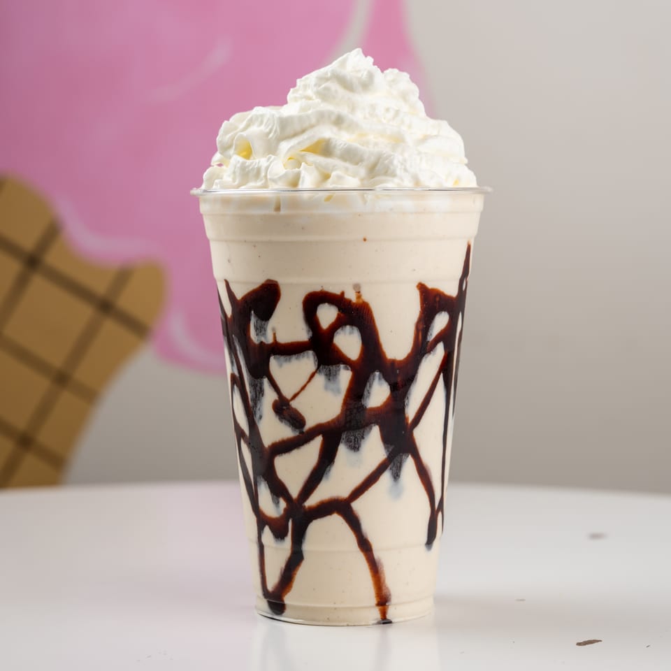 Creamy chocolate-drizzled milkshake topped with whipped cream in a clear cup, set against a pink and gray backdrop.