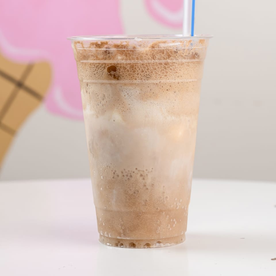 Clear plastic cup of frothy iced coffee with a blue straw on a white table against a pink and beige background.