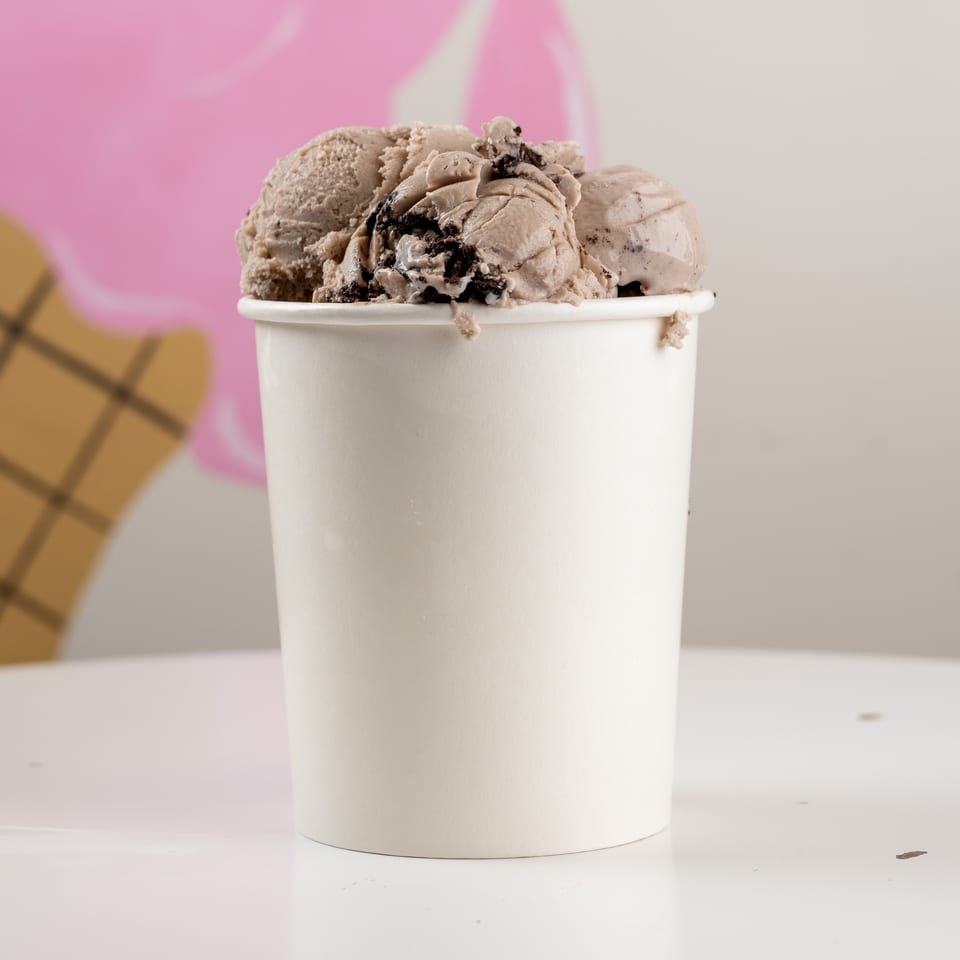 White cup overflowing with scoops of chocolate ice cream and cookie pieces, against a pink and beige backdrop.