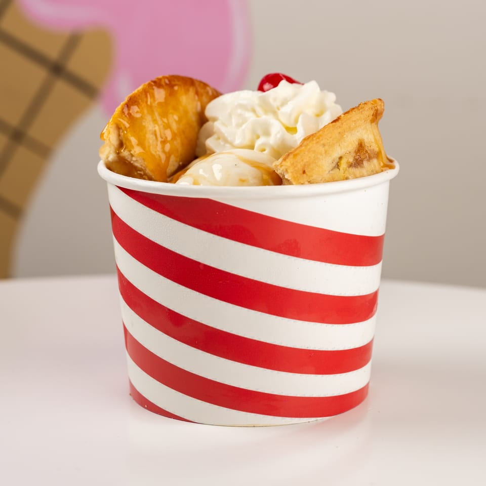 Red-and-white striped cup filled with ice cream, whipped cream, caramel drizzle, cherries, and pastry pieces.