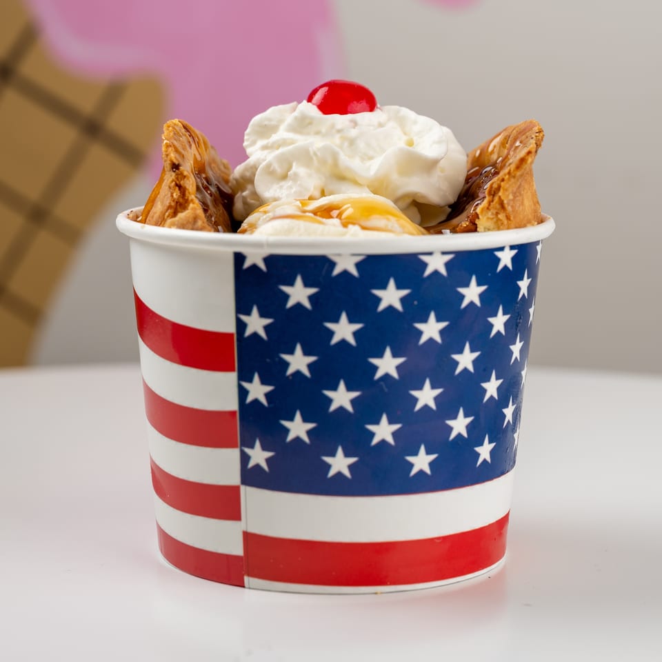 Vanilla ice cream sundae topped with whipped cream, cherry, caramel, and pastry pieces in patriotic red, white, and blue cups.
